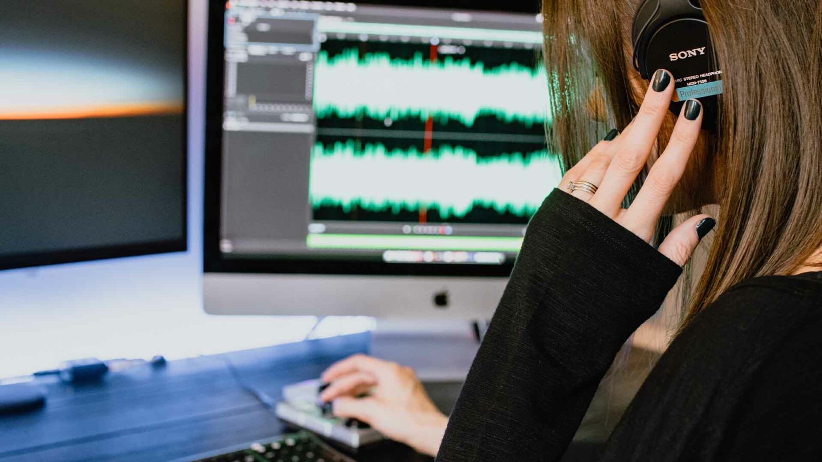 O persoană folosește căști profesionale Sony mc stereo headphon mdr-7500 pentru a asculta ieșirea multimedia de la computerul personal, care este conectat la un monitor de computer și la alte dispozitive electronice într-un cadru interior. Folosind un schimbător de voce ai.
