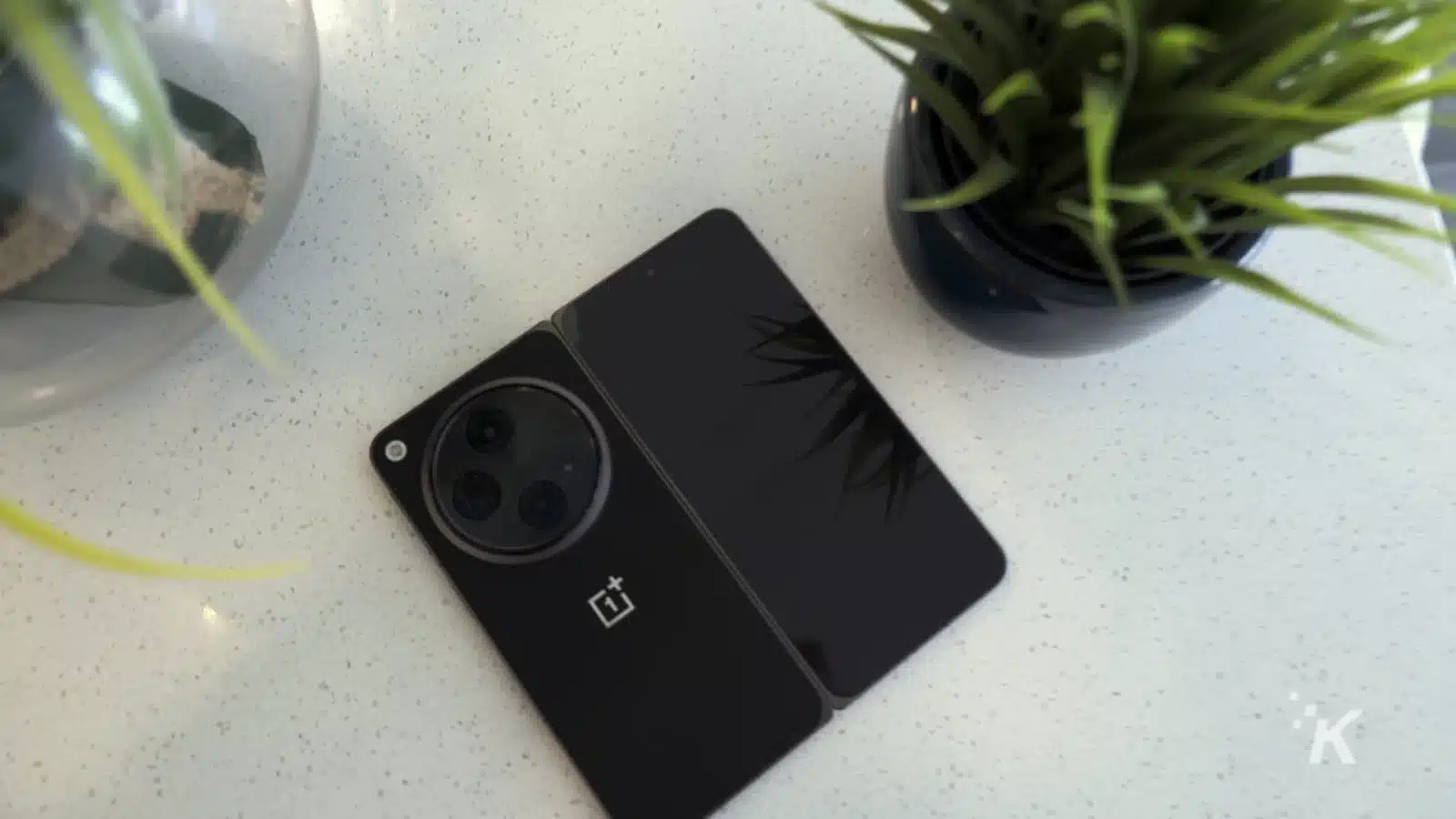 ulasan: oneplus terbuka A houseplant blooms in a flowerpot next to a gadget and mobile phone on an indoor table.