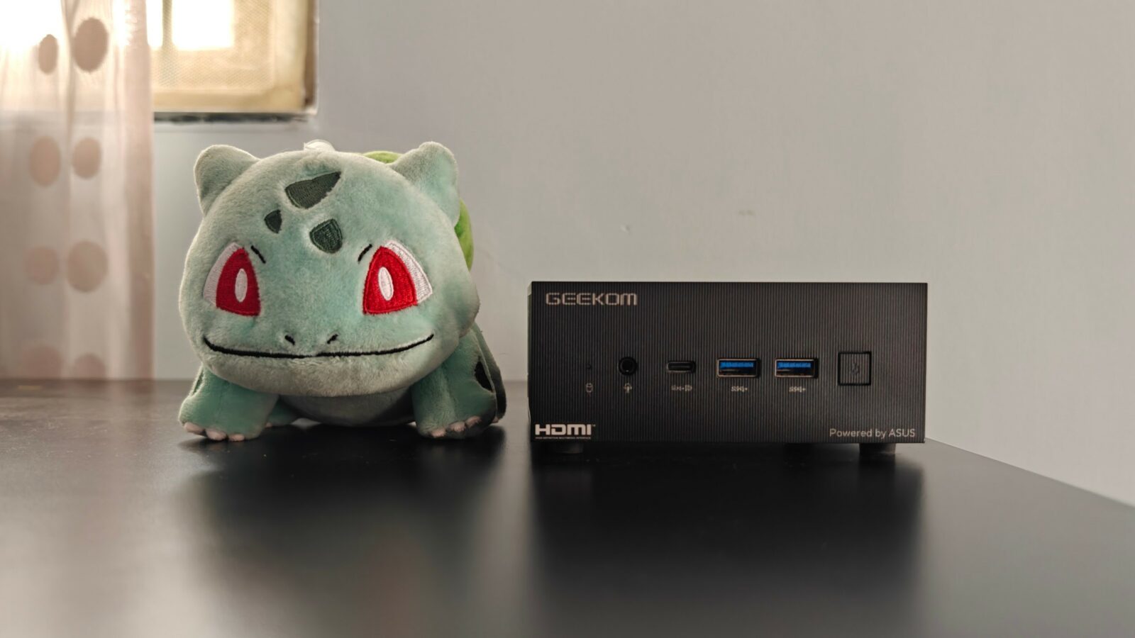 ตุ๊กตา Bulbasaur วางอยู่ข้างๆ มินิพีซีเกินบรรยายบนพื้นผิวสะท้อนแสง โดยมีผ้าม่านและหน้าต่างเป็นพื้นหลัง