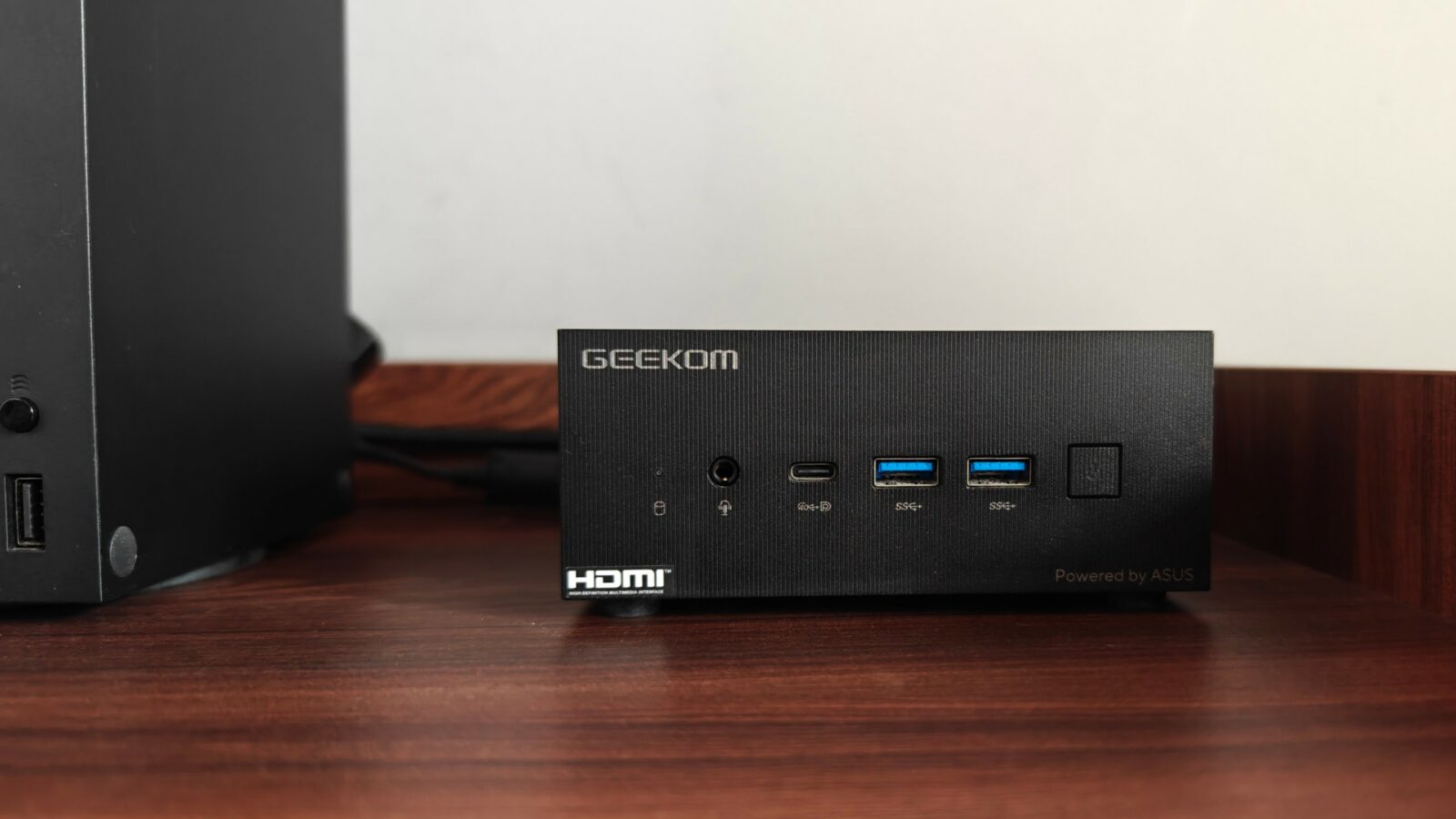 มินิพีซีสีดำขนาดเล็กที่มีพอร์ตหลายพอร์ตที่มีป้ายกำกับว่า "geekom" บนพื้นผิวไม้ ติดกับหอคอมพิวเตอร์ขนาดใหญ่ โดยมีผนังสีขาวเป็นฉากหลัง