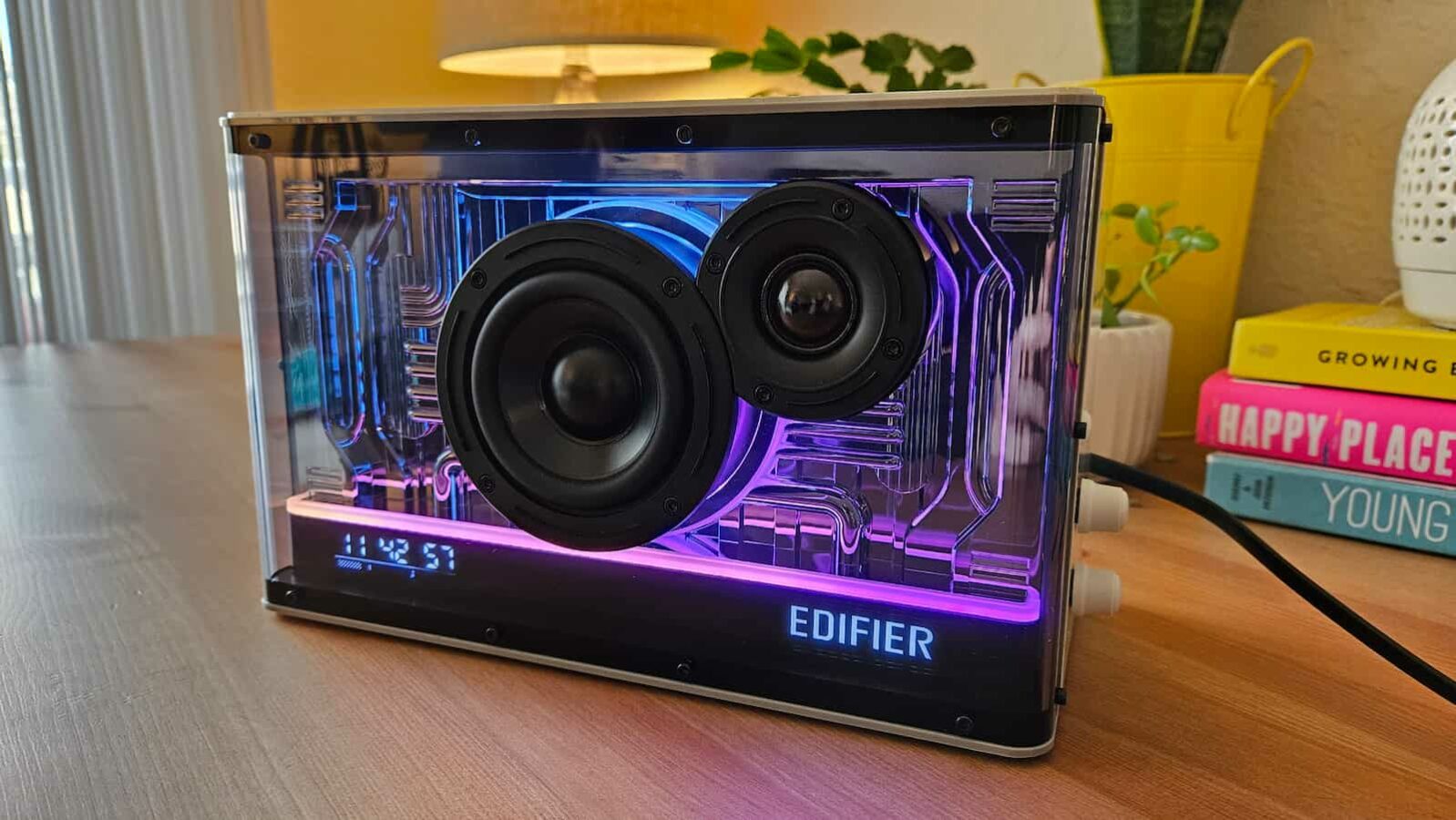 ลำโพงโปร่งใส edifier พร้อมไฟส่องสว่างภายในสีม่วงและสีน้ำเงินแสดงอยู่บนพื้นผิวไม้ โดยมีหนังสือและของตกแต่งเป็นพื้นหลัง