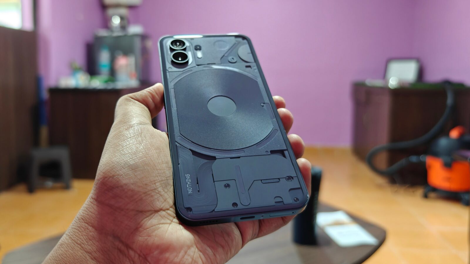 มือคนถือสมาร์ทโฟนพร้อมเคสใสเผยให้เห็นรายละเอียดภายใน พื้นหลังเป็นห้องเบลอๆ มีผนังสีม่วงและเฟอร์นิเจอร์