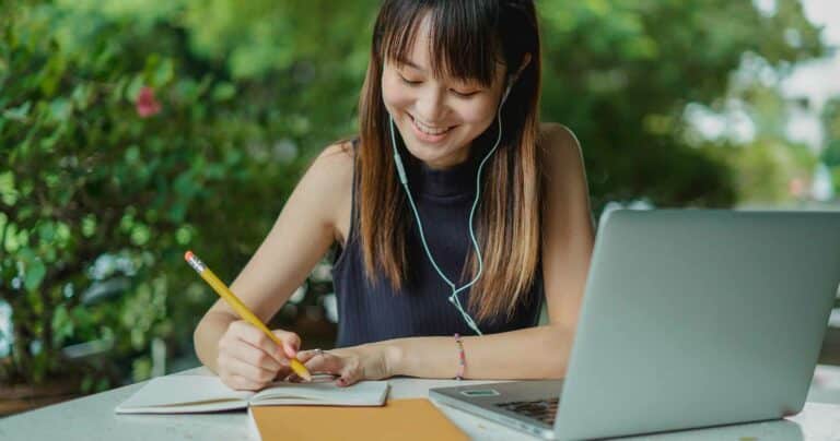 Woman writing on notepad with a laptop next to her.