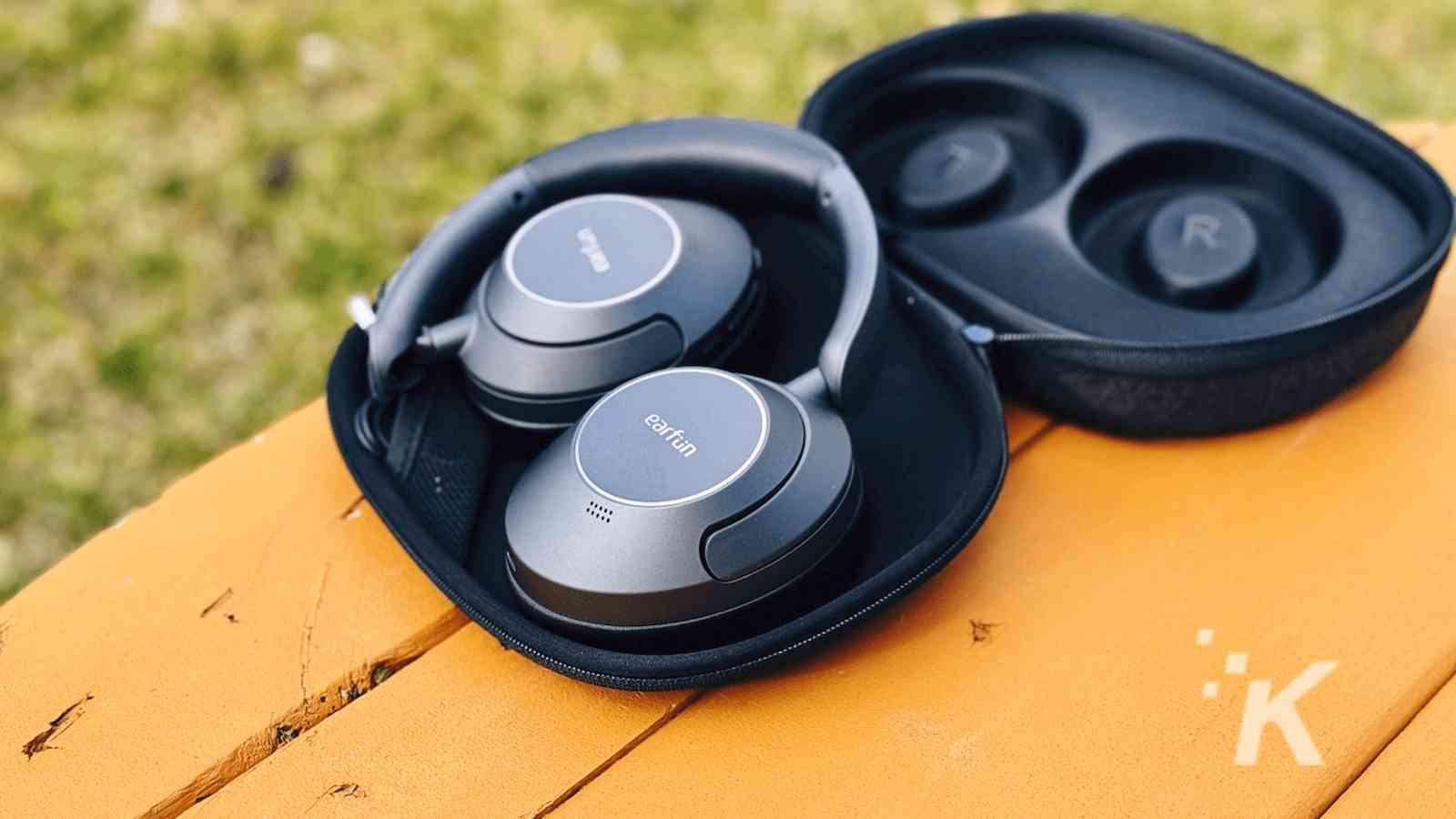 A pair of black over-ear headphones placed partially inside an open, black carrying case on a yellow wooden bench.